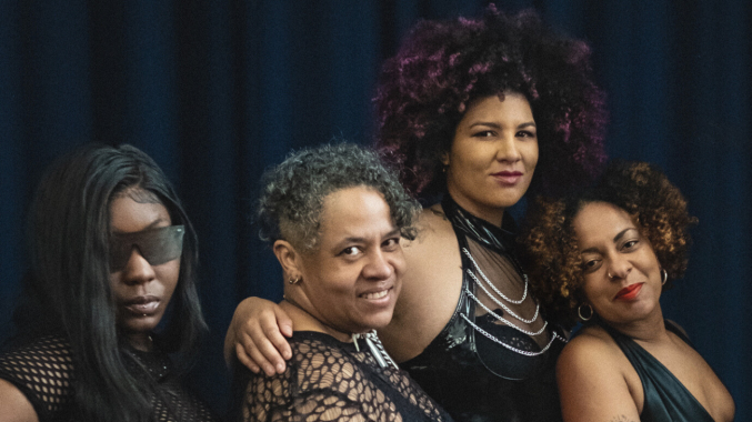 Four black women posing in a row with a black fabric backdrop behind them