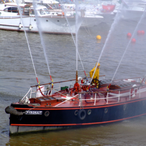 Pyronaut, a navy and red fire boat, on the water. There is one crew member on board wearing all yellow. The boat is shooting water in different directions out of several hoses. 