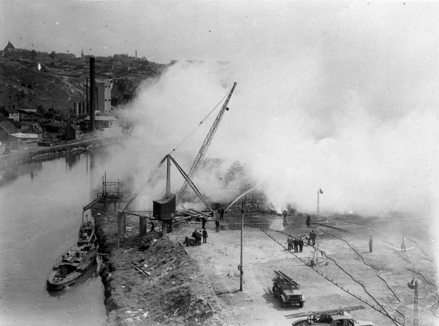 A scene of the docks in the early 1940's. Pyronaut sits in the water next to a crane, behind the crane is a large cloud of smoke obscuring most of the scene. 